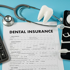 Dental insurance form on blue surface next to calculator, model tooth, stethoscope, and other dental supplies